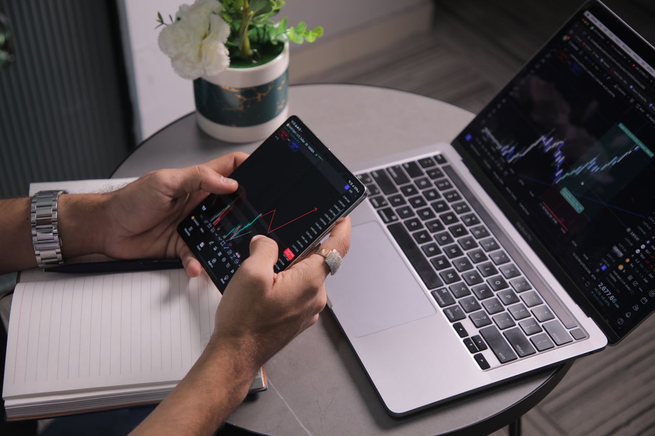 Un homme analyse les tendances boursières sur un espace de travail moderne, avec un ordinateur portable et une application de trading sur mobile.